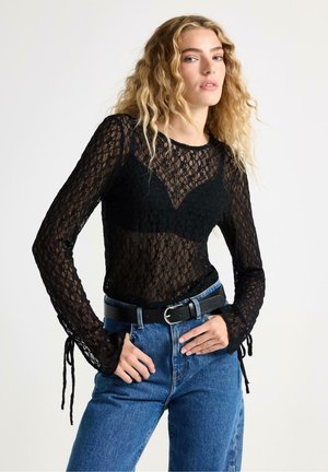 Musta pitsinen pitkähiha tunika, jossa on tiukka vartalo-osa, läpikuultava rakenne ja solmukohtia ranteissa, yhdistettynä korkeavyötäröisiin sinisiin farkkuihin ja mustaan vyöhön.