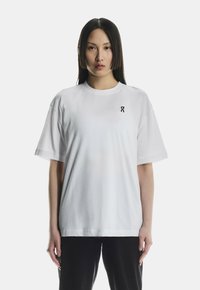 Kvinde med glat, mørkt hår iført en løs hvid t-shirt med et lille sort logo på venstre bryst og sorte bukser, der står mod en hvid baggrund.