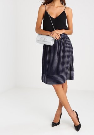 Kobieta ubrana w czarny bezrękawnik, granatową spódnicę do kolan w paski, czarne szpilki, trzymająca małą srebrną torebkę na łańcuszku.