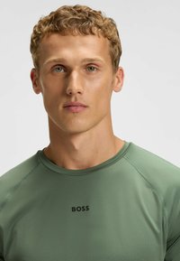 Grønn atletisk t-skjorte laget av et glatt stoff, med rund hals og "BOSS"-logoen i svart i midten.