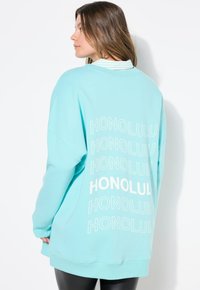 Sudadera de gran tamaño en color verde menta con el texto "HONOLULU" en blanco en la parte de atrás. Mangas largas con puños acanalados, textura de tela suave.