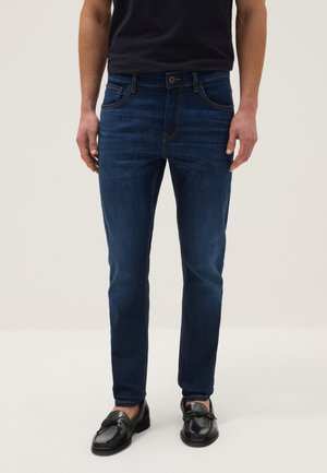 Hombre con jeans ajustados azul oscuro y mocasines de cuero negros, de pie contra un fondo liso y claro.