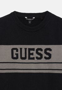 Svart strikkegenser med en grå horisontal stripe med "GUESS" i fete sorte bokstaver. Ribbestrikket rund hals og teksturert stoff.