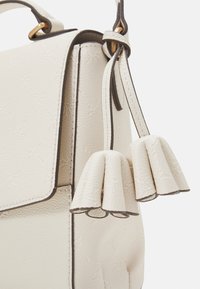 Valkoinen nahkalaukku, jossa on teksturoidun pinnan lisäksi läppäsuljin ja koristeelliset tupsut. Vankka olkahihna kontrastivärissä.