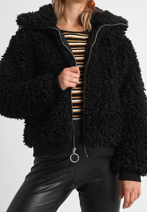 Femme portant une veste noire texturée zippée sur un haut rayé noir, blanc et jaune, avec un pantalon en cuir noir.