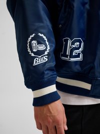 Manche de veste bleu marine avec logo de couronne de laurier blanche représentant une chope de bière et un hotdog, texte "BEE'S" et numéro "12" sur le côté.