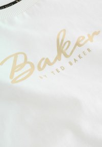 Tela de algodón blanca con una gran impresión dorada brillante de la palabra "Baker" y "by Ted Baker" en un texto más pequeño debajo.
