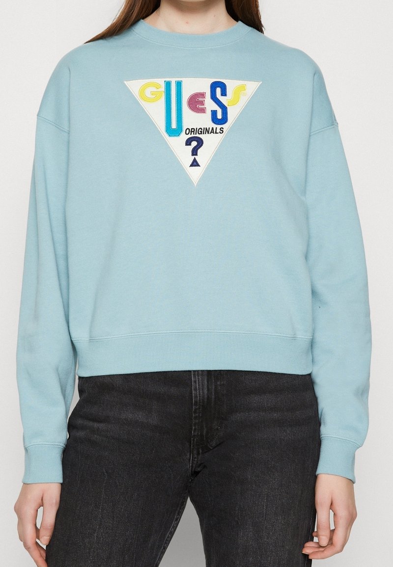 Ljusblå sweatshirt med ribbade cuffs och nederkant, med en triangulär broderad grafik med "GUESS" och "ORIGINALS" i olika färger.