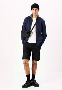 Chaqueta con cremallera azul marino, camiseta blanca, pantalones cortos negros, calcetines negros y zapatos. Bolso cruzado negro. El modelo está de pie contra un fondo sencillo.
