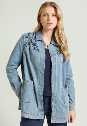 Mujer con cabello largo y rubio que lleva una chaqueta de mezclilla azul claro con bordados florales en los hombros y bolsillos delanteros con parche.