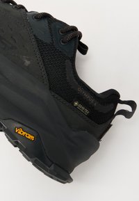 Czarne buty trekkingowe z teksturowaną siateczką górną, podeszwą Vibram, logo GORE-TEX i wyraźnymi sznurowadłami. Charakteryzują się masywną konstrukcją i gumowymi akcentami.