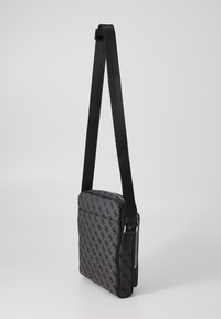 Bolso de mensajero negro con tejido estampado, cierre de cremallera y una correa ajustable. Presenta un bolsillo frontal plano y forma rectangular.