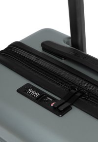 Valigia rigida grigia con superficie testurizzata, dotata di una zip nera e un lucchetto a combinazione. Manico estraibile per una facile mobilità.