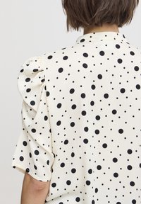Short-sleeve blouse in cream with black polka dots, featuring puffed sleeves and a classic collar. Smooth fabric with a soft texture.