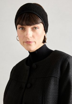 Czarna teksturowana opaska na głowę w połączeniu z czarną bluzką z wysokim dekoltem i okrągłymi guzikami, na neutralnym tle.