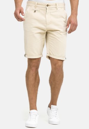 Hombre con pantalones cortos beige remangados y zapatillas blancas, de pie contra un fondo liso y claro con las manos a los lados.