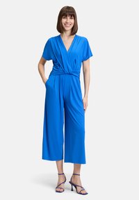 Donna sorridente che indossa una tuta corta manica corta di un luminoso blu con gambe ampie e sandali con tacco alto blu coordinati.