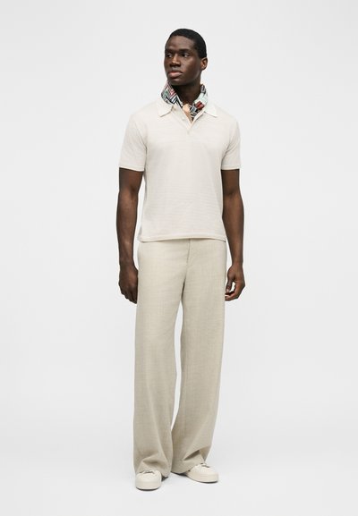 Άνδρας που φοράει μπεζ πόλο με κοντά μανίκια, φαρδιά μπεζ παντελόνια, λευκά αθλητικά παπούτσια και ένα κασκόλ με σχέδια γύρω από τον λαιμό, στέκεται μπροστά σε απλό φόντο.