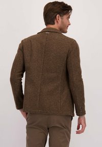 Man die wegkijkt, gekleed in een bruine wollen blazer en bijpassende bruine broek tegen een effen lichte achtergrond.