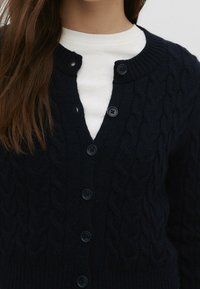 Mörkblå stickad cardigan med kabeldesign, rund halsringning och svarta knappar. Över en vit topp.