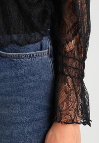 Blusa nera in pizzo con maniche lunghe trasparenti, caratterizzata da intricati motivi floreali e bordi scallop, abbinata a jeans in denim blu a vita alta.