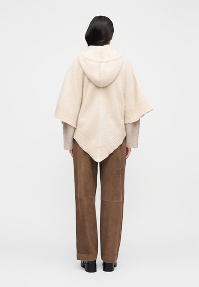 Poncho beige à capuche avec bords côtelés, en matériau doux. Associé à un pantalon marron à jambes larges et des chaussures noires. Fond uni.