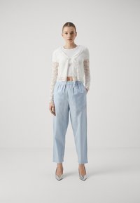 White lace cardigan, cropped design, tied in front. Light blue elastic-waist trousers, loose fit. Silver pointed heels. Simple background.