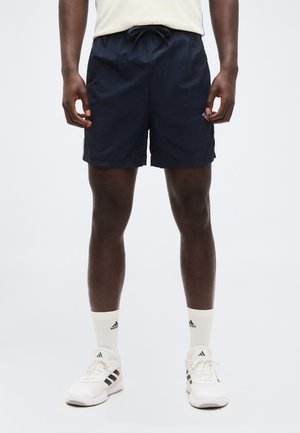 Uomo con pantaloncini sportivi blu navy, calzini bianchi Adidas e scarpe bianche Adidas, in piedi davanti a uno sfondo chiaro e uniforme.