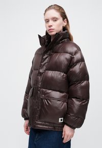 Chaqueta acolchada marrón con acabado brillante, que presenta un cuello alto, cierres de botón y un bolsillo en la parte frontal. Corte relajado con puños elásticos.