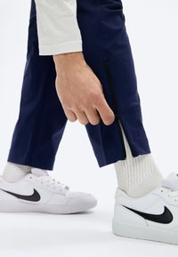 Άτομο που φοράει μπλε παντελόνι με πλευρικό φερμουάρ και το διορθώνει, άσπρες κάλτσες και άσπρα αθλητικά παπούτσια Nike με μαύρο λογότυπο swoosh.