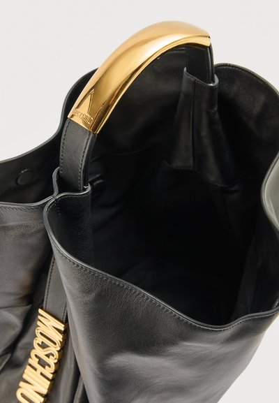 Sac cabas en cuir noir avec accent de poignée dorée et pendentif logo Moschino suspendu à la sangle, vu de dessus.