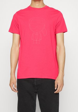 Kirkkaan punainen T-paita, jossa on hienovarainen kohokuvioitu sarjakuvahahmo aurinkolaseissa, solmiossa ja poninhäntä, yhdistetty mustiin housuihin.