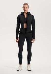 Černá sportovní bunda s přiléhavým střihem a kapucí, vrstvená přes černý crop top a legíny s vysokým pasem, doplněná o světlé tenisky.