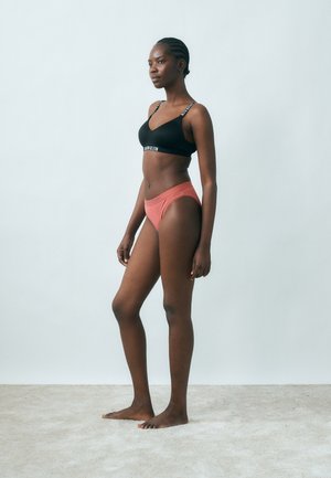 Νεαρή γυναίκα που στέκεται ξυπόλυτη σε καλυμμένο με χαλί πάτωμα, φορώντας μαύρο σουτιέν και κοραλί εσώρουχο, στραμμένη προς τα αριστερά μπροστά σε απλή ανοιχτόχρωμη τοίχο.