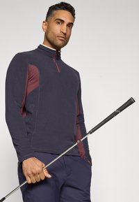 Pullover blu scuro a tre quarti con zip, caratterizzato da pannelli laterali bordeaux e dettagli testurizzati. Il modello tiene una mazza da golf, indossando pantaloni blu navy.