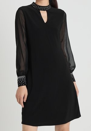 Robe noire à manches longues transparentes, petite ouverture en forme de trou de serrure à l'avant, col et poignets cloutés sur une personne debout devant un fond neutre.