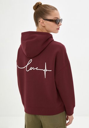 Vrouw met haar in een knot, met schildpaddenbril en een bordeaux hoodie met "love" en een hartslagontwerp op de rug, van opzij gezien.