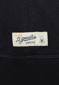 Tela negra mostrando una etiqueta de ropa con el texto "D. Gaastra" en cursiva, rodeada por un fondo beige. Detalle de costura visible.