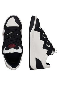 Black and white sneakers with textured black accents, featuring a round toe, flat sole, and black laces. Red logo detail on tongue.