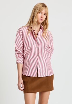 Jeune femme aux longs cheveux blonds portant une chemise à rayures rouges et blanches boutonnée et une mini-jupe marron, sur un fond uni.