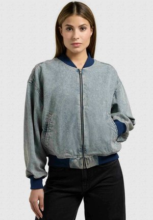 Mladá žena v světlé modré džínové bomber bundě s námořnicky modrými manžetami, límcem a spodním lemem, v černých kalhotách, stojící vzpřímeně.