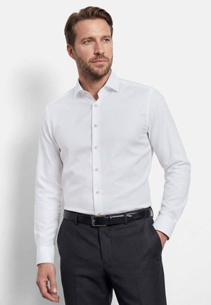 Mann, der ein eng anliegendes weißes Hemd, dunkelgraue Hose und schwarzen Gürtel trägt, steht mit einer Hand in der Tasche vor weißem Hintergrund.