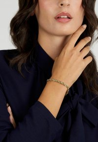 Bracciale d'oro intrecciato su un polso, che mostra un design a treccia. Chi lo indossa ha una mano posata leggermente sul volto, con una blusa scura.