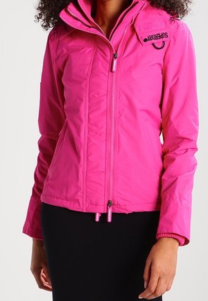 Femme portant une veste zippée rose vif avec un col montant et un écusson logo, associée à une jupe noire, sur un fond uni.