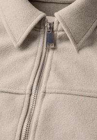 Chaqueta de mezcla de lana gris claro con un cuello doblado y un cierre de cremallera plateado de longitud completa, con costuras visibles y una textura suave.