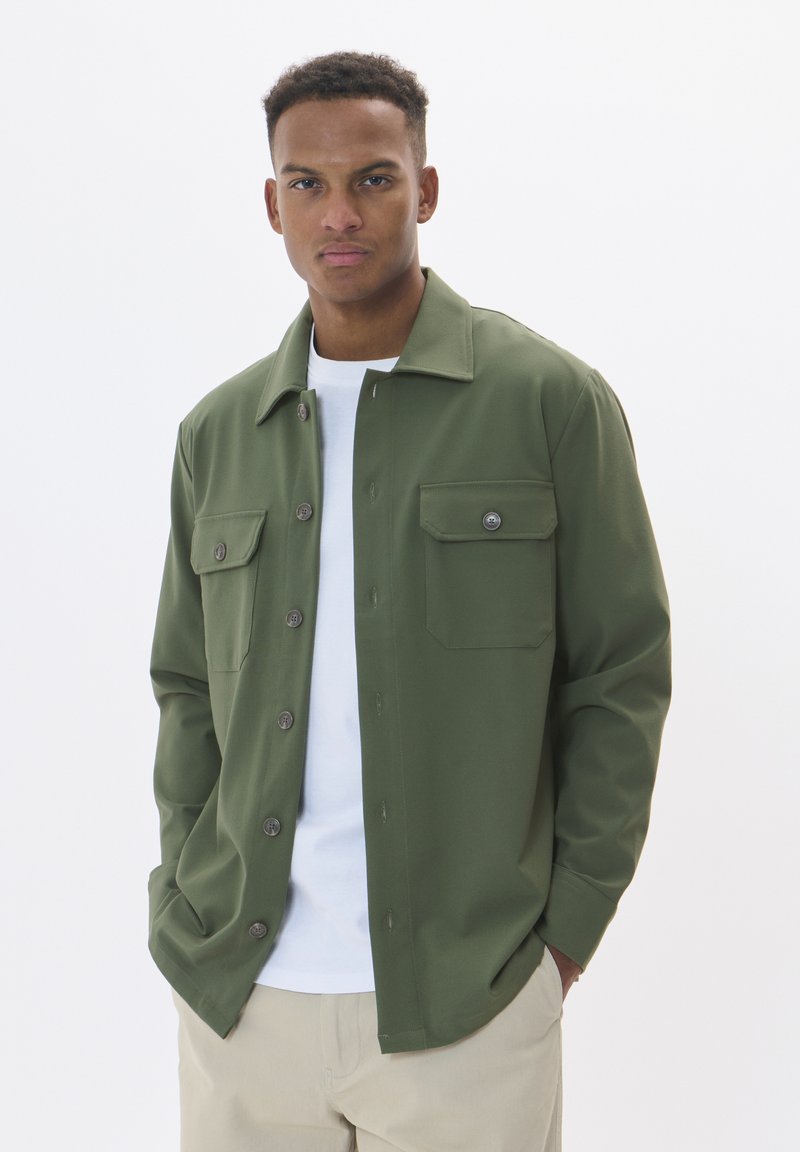Olivengroen button-up shirt van soepel stof, met twee borstzakken, een spread collar en contrasterende knopen. Draagt over een wit t-shirt.