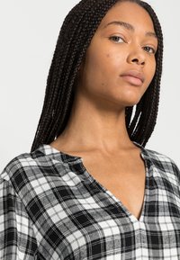 Camisa de xadrez em preto e branco com decote em V, padrões de xadrez contrastantes e mangas curtas, feita de um tecido suave com textura visível.