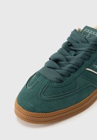 Žalias zomšiniai sportbačiai su tekstūruotu audiniu, plokščiomis raištelėmis ir smėlio spalvos guminiu padu. Šone matomas logotipas; elegantiška, apvali forma.