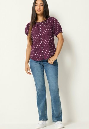 Vrouw met lang, steil haar die een paarse blouse met korte mouwen en knopen draagt met witte stippen, een blauwe spijkerbroek en witte sneakers.