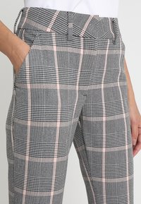 Close-up van een persoon die een hoge taille geruite broek draagt met zwart, wit en lichtroze ruiten, en één hand ontspannen in de zak heeft.